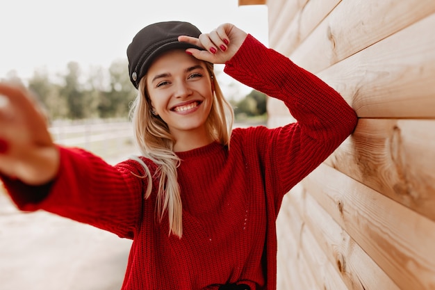 Charming blonde in red pullover and dark hat happily smiling at her phone. Pretty girl making selfie near wooden house outdoor.