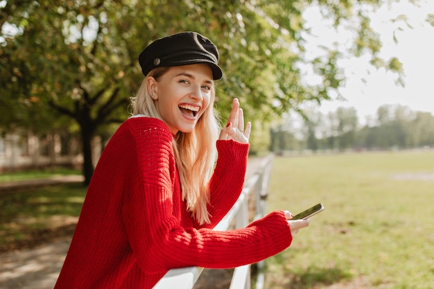 Очаровательная блондинка счастливо улыбается, держа телефон. Сияющая девушка, имеющая хороший день в солнечном парке.