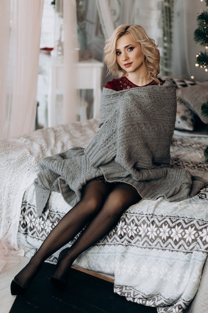 Charming blonde woman envelopes herself in grey plaid sitting on a bed before a Christmas tree