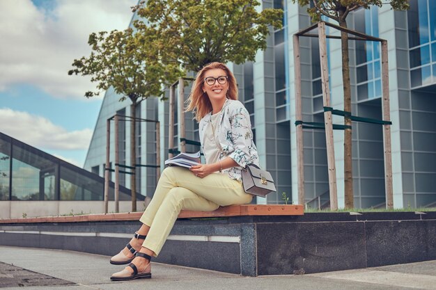 현대적인 옷을 입은 매력적인 금발 여성, 책을 가지고 공부하고, 고층 빌딩을 배경으로 공원 벤치에 앉아