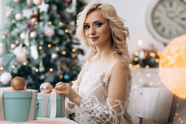 Charming blonde woman opens present boxes sitting before a Christmas tree