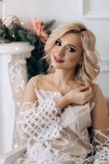 Charming blonde woman in white dress poses in a room with large Christmas tree