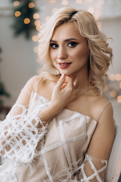 Charming blonde woman in white dress poses in a room with large Christmas tree