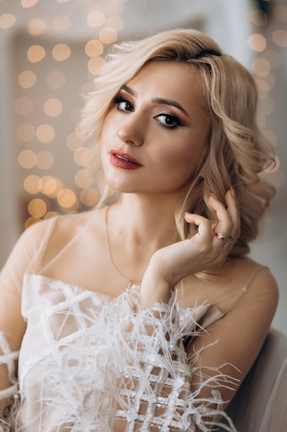 Charming blonde woman in white dress poses in a room with large Christmas tree