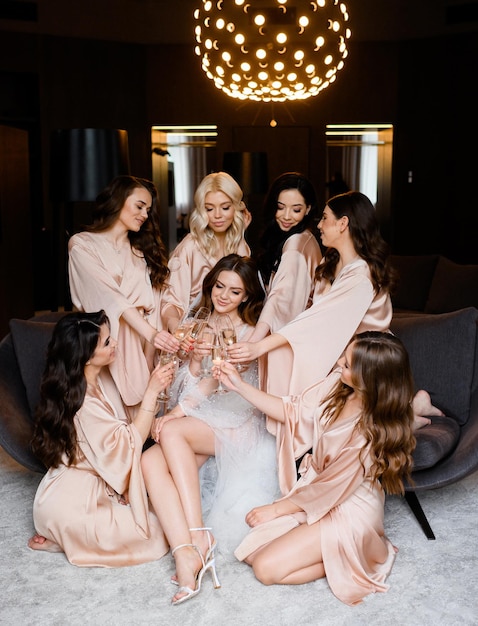 Charming bride in elegant wedding dress celebrating wedding day clinking glasses with champagne with bridesmaids Beutiful girls friends of bride in satin robe bachelorette party in luxury apartment