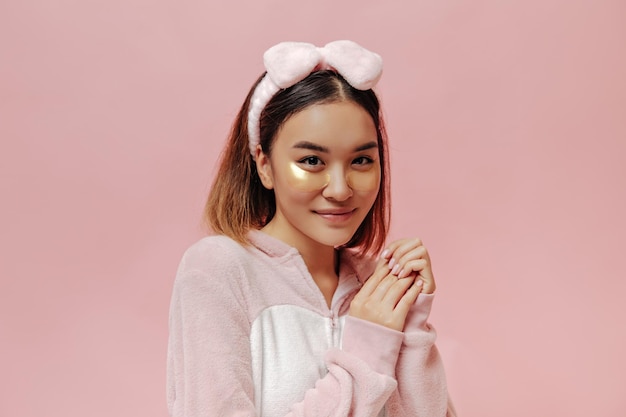 Charming browneyed woman in headband and kigurumi smiles and poses with eyepatches on pink background