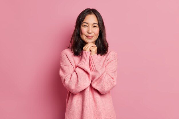 Charming brunette Asian girl with tender expression has rouge cheeks keeps hands under chin dressed in loose jumper poses 