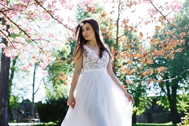 Charming brunette bride walks in white dress among blooming sakura trees