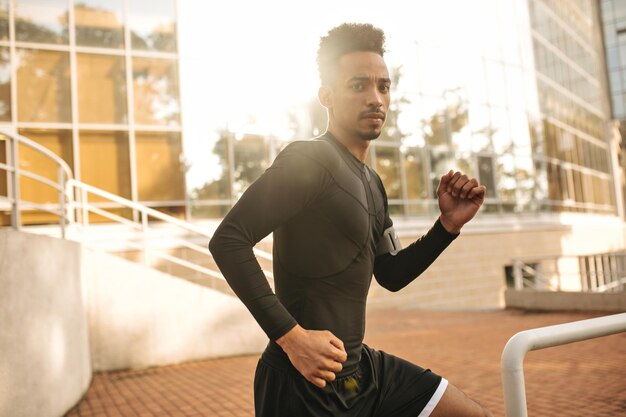 Charming brunette darkskinned man in sport black outfit running outside Young guy looking into camera and working out