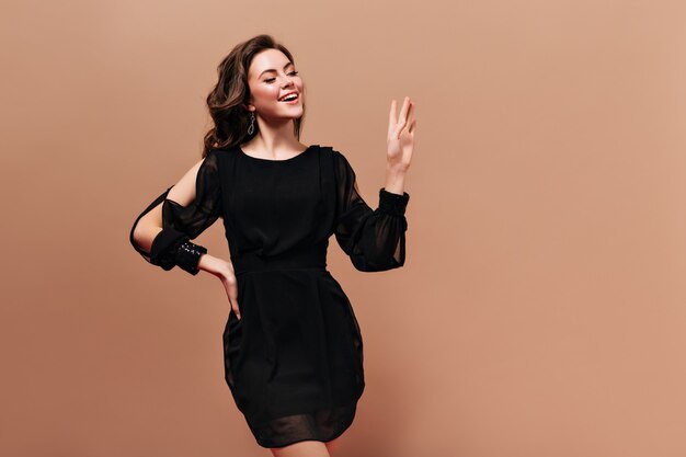 Charming brunette girl in black dress is smiling and waving hand in greeting on beige background.