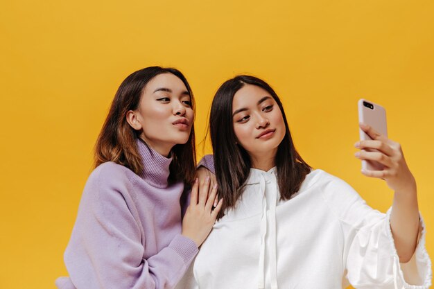 Charming brunette girls in sweatshirts take selfie on isolated Attractive young woman in white hoodie holds phone on orange background