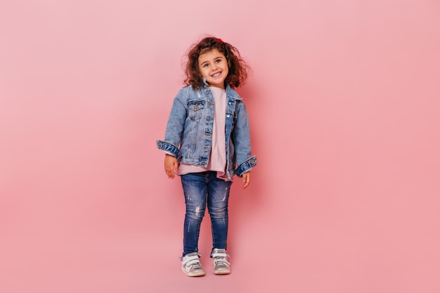 Free photo charming brunette kid in denim attire smiling at camera. full length view of happy preteen girl isolated on pink background.