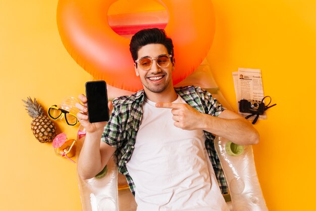 Charming brunette man in plaid shirt and orange glasses points to black smartphone. Guy is resting on inflatable mattress.
