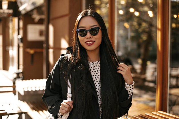 Charming brunette woman in black trench coat and sunglasses smiles widely and holds handbag Asian lady walks outside