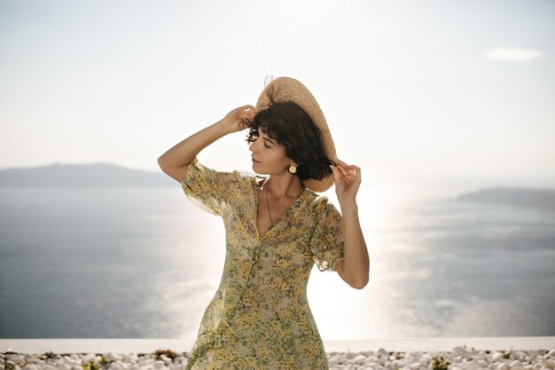 보트와 꽃무늬 드레스를 입은 매력적인 브루네트 여성은 바다 배경에서 포즈를 취합니다. 곱슬거리는 여성은 모자를 들고 화창한 여름날 야외에서 즐깁니다.