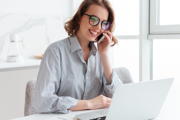 Charming brunette woman in glasses and casual wear talking on mobile phone while sitting at table and looking at laptop screen