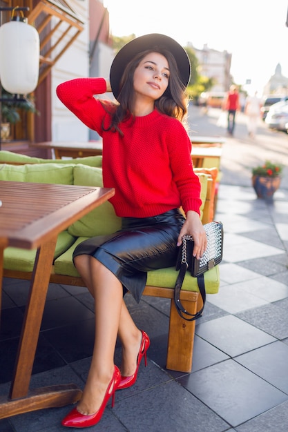 Charming brunette woman in red autumn knitted sweater and leather skirt relaxing on sofa in open space restaurant.