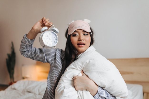 Очаровательная брюнетка в полосатой пижаме смотрит на будильник и обнимает белую подушку