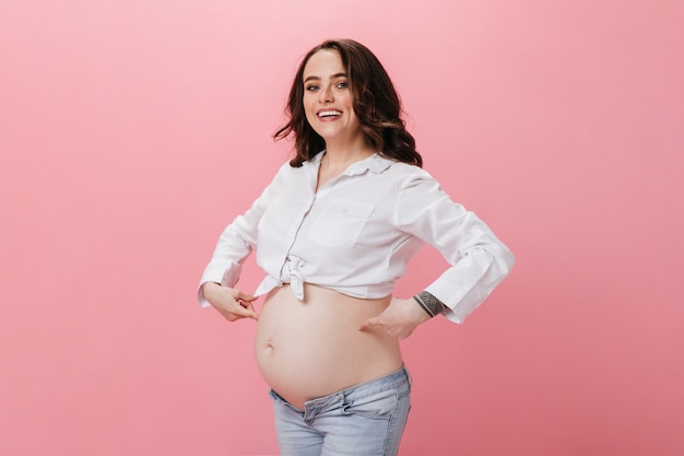 Charming brunette woman in white shirt and jeans points at her belly. Pregnant lady smiles widely on pink isolated background.