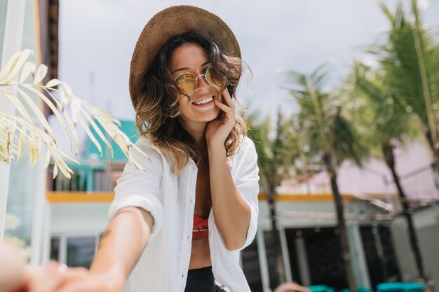 Charming brunette woman in white shirt making selfie with palm trees.