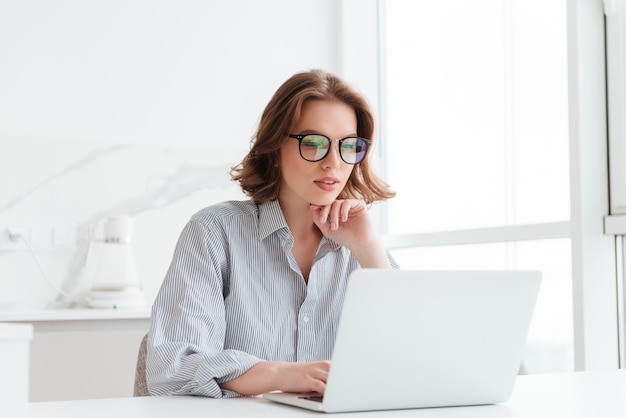 メガネとストライプシャツの魅力的な女性実業家が自宅に座ってラップトップコンピューターでの作業