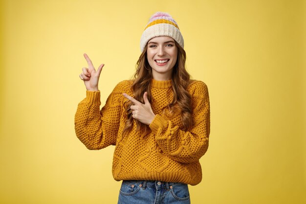 왼쪽 위 모서리를 옆으로 가리키는 모자 스웨터를 입은 매력적인 20대 젊은 여성이 흥미로운 링크를 공유하고 멋진 프로모션을 기뻐하며 멋진 제품, 노란색 배경을 추천합니다.