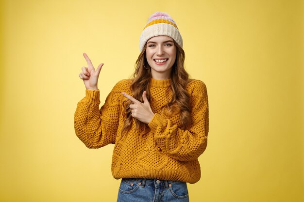 Charming carefree smiling young 20s woman wearing hat sweater pointing upper left corner sideways sharing interesting link awesome promotion delighted recommend cool product, yellow background