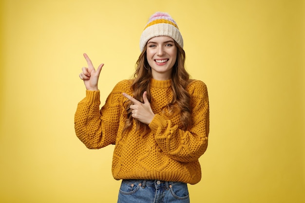 Charming carefree smiling young 20s woman wearing hat sweater pointing upper left corner sideways sharing interesting link awesome promotion delighted recommend cool product, yellow background