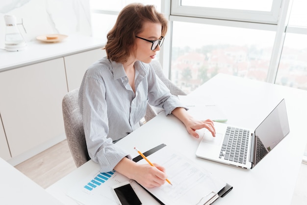 charming caucasian businesswoman in striped shirt working with laptop and papers in light apartment