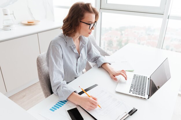 charming caucasian businesswoman in striped shirt working with laptop and papers in light apartment