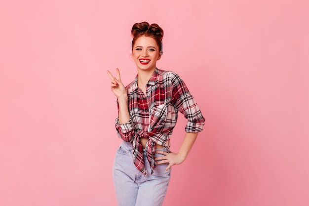 Charming caucasian lady in checkered shirt showing peace sign. Studio shot of smiling pinup woman isolated on pink space.