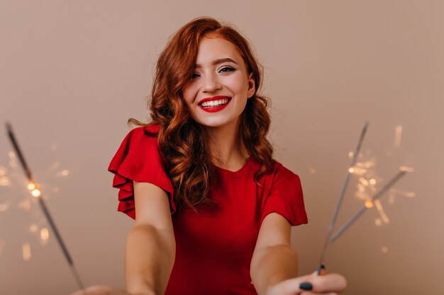 Charming caucasian woman in red dress holding bengal lights. Adorable girl celebrating new year with smile.