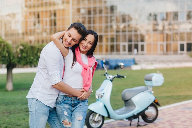 Free photo charming caucasian woman with pink sweater embracing husband during photoshoot in park. happy married young couple posing beside scooter on blur city background.