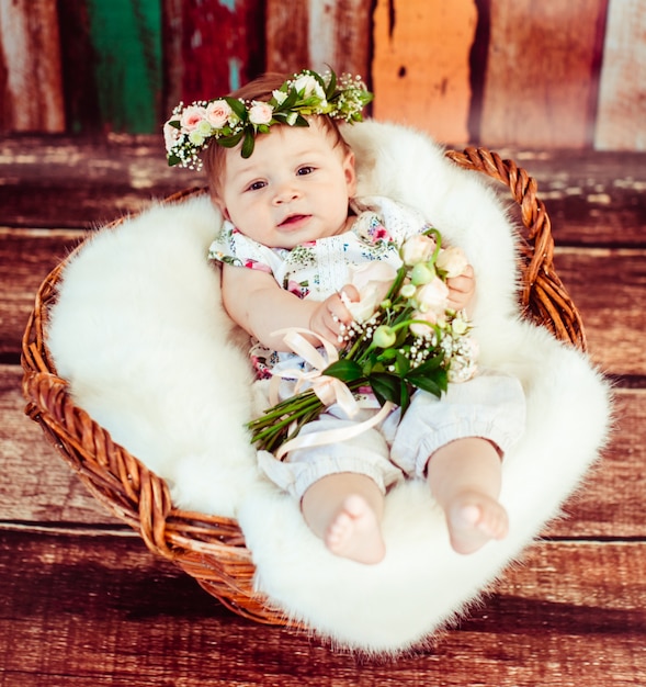 Charming child in rose wreath lies on fluffy blanket in brown basket