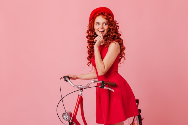 ピンク色の空間で自転車でポーズをとる、気分のいい魅力的なコケティッシュな女性が横にずる賢く見えます。