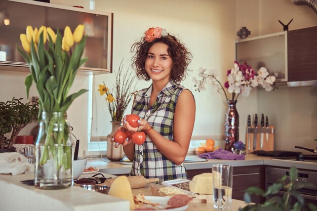 Charming curly Hispanic girl holds fresh tomatoes while cooking in her kitchen.