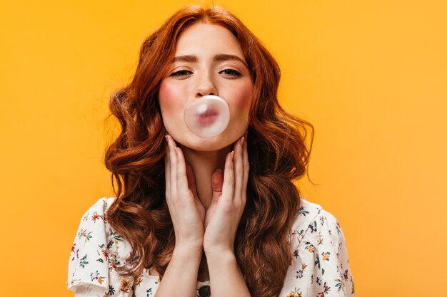 Charming curly red-haired woman in white top makes bubble of gum. Portrait of young woman on orange background.