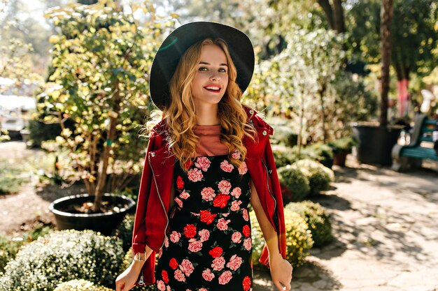 Charming curly woman in wide-brimmed hat and dress with roses with smile posing in park.