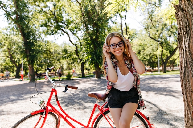 公園で音楽を聴いている夏の服装の魅力的なかわいい女の子。自転車の横に立っているヘッドフォンで陽気な金髪モデルの屋外写真。