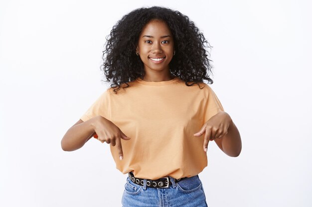 魅力的なかわいい活気のある若いアフリカ系アメリカ人の20代の女性アフロヘアカット笑顔フレンドリーな人差し指を下に向けて興味深い提案、プロモーションアクション、立っている白い壁を示しています
