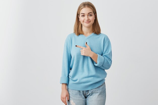 Charming cute young European female customer smiling and pointing index finger sideways, showing blank white wall with copy space for your promotional content