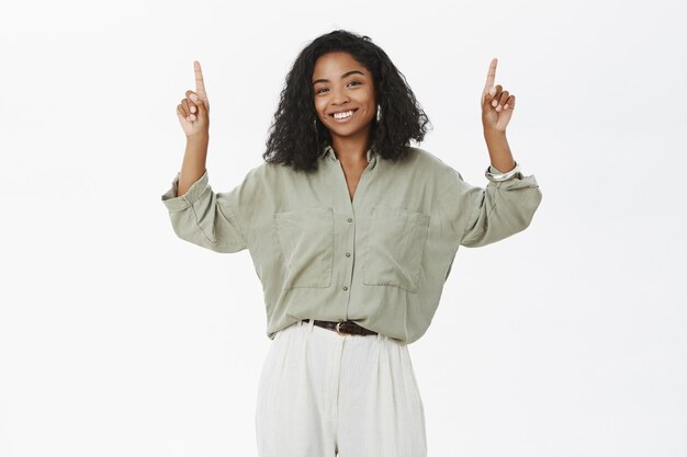 Charming dark-skinned female model with curly hairstyle in trendy blouse and pants raising arms and pointing up