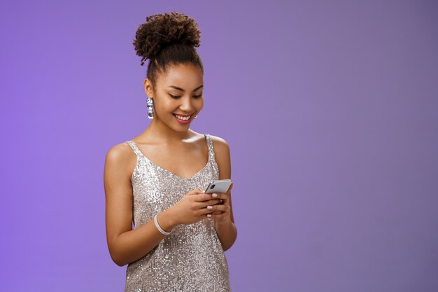 Charming elegant african-american woman in stylish glittering prom dress writing message calling friend hold smartphone look amused happy smile phone display using gadget app scroll party photos.