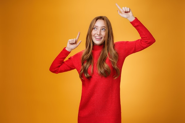 Charming and elegant attractive redhead female in red knitted dress raising hands up carefree as dancing having fun being pleased and happy gazing amused with broad grin at upper left corner