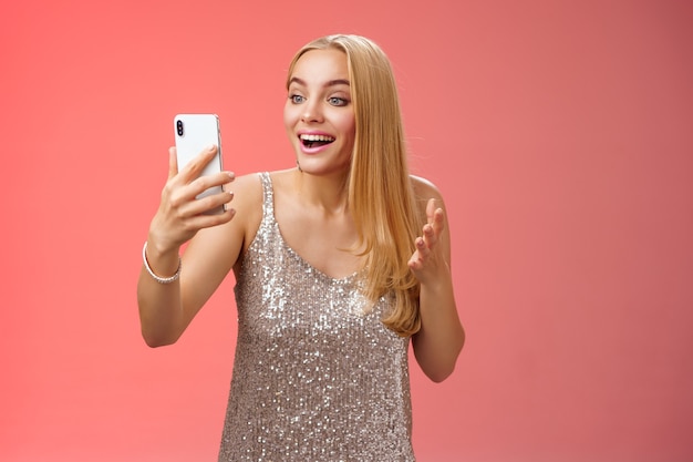 Charming elegant nice blond girl in silver dress talking video call speaking looking smartphone display amused surprised smiling happily have conversation sibling showing prom outfit