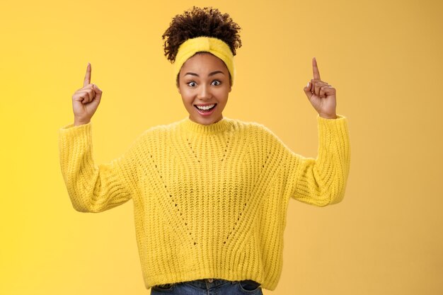 Charming emotive young african-american woman in sweater headband winning impressive sum money pointing up index fingers showing link astonished surprised smiling broadly yellow background.