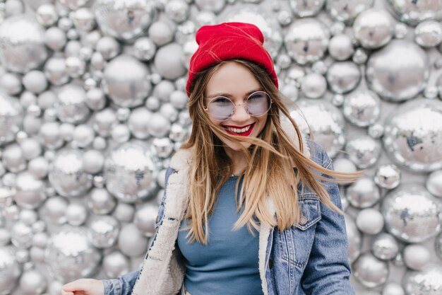 행복 한 미소와 함께 포즈를 취하는 귀여운 빨간 모자에 매력적인 유럽 소녀. 스파클 디스코 공 옆에 서있는 데님 재킷에 예쁜 금발의 여자의 사진.