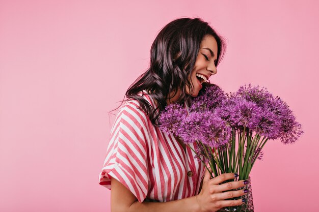 Charming European girl with beautiful tan poses with armful of lilac tsatov. Closeup portrait of lady in white and pink top.