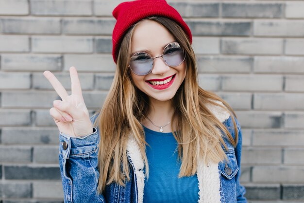 Charming european woman in casual clothes posing with cute smile and peace sign. Outdoor shot of elegant laughing girl in red hat.