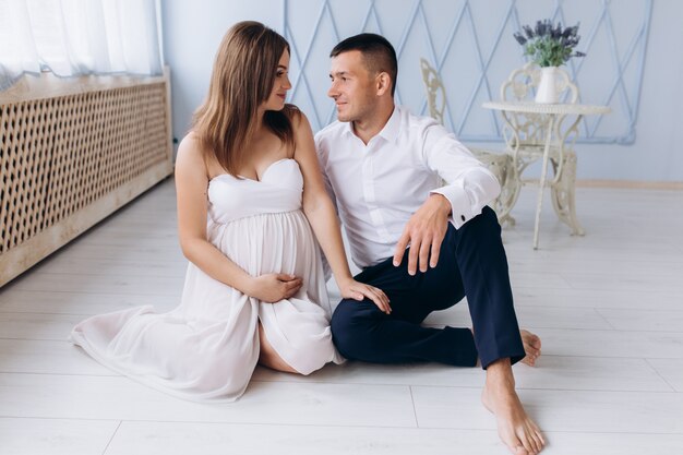 Charming expecting couple sits on a floor in a bright white room